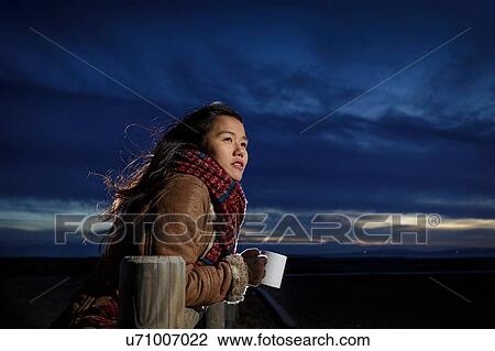 Stock Image - Young woman wrapped in scarf looking out from beach at dusk, Tarragona, Catalonia, Spain. Fotosearch
