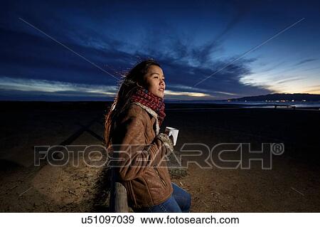 Stock Photo - Young woman wrapped in scarf looking out from beach at dusk, Tarragona, Catalonia, Spain. Fotosearch