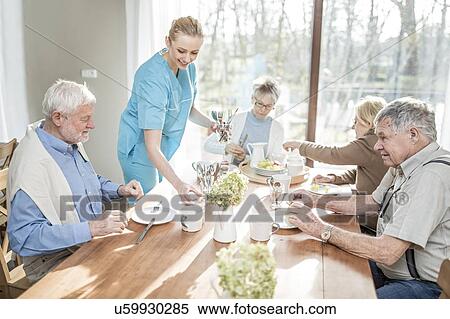 Stock Photography - Female care worker serving senior adults at dinner table in care home.. Fotosearch