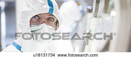 Lab technician wearing a protective suit, face mask and safety goggles in a laboratory that must maintain a sterile environment. View Large Photo Image Picture - Lab technician wearing a protective suit, face mask and safety goggles in a laboratory that must maintain a sterile environment.. Fotosearch