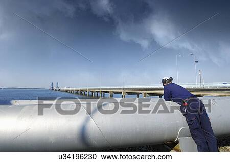 Stock Image - Oil and gas worker leaning on giant pipelines leading out to sea.. Fotosearch