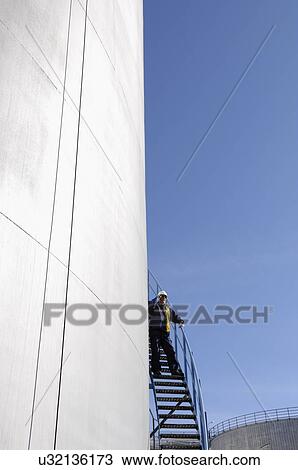 Stock Image - Refinery worker standing on ladder talking on phone next to industrial oil and fuel storage tanks.. Fotosearch