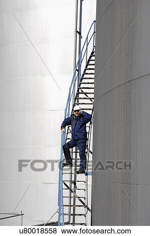 Stock Photo - Refinery worker standing on ladder talking on phone next to industrial oil and fuel storage tanks.. Fotosearch