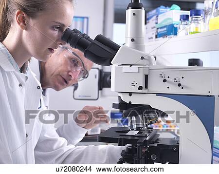 Scientists viewing a sample on a glass slide under a light microscope. View Large Photo Image Picture - Scientists viewing a sample on a glass slide under a light microscope.. Fotosearch