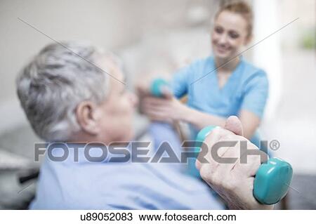 Stock Image - Senior man holding hand weight, physiotherapist helping.. Fotosearch