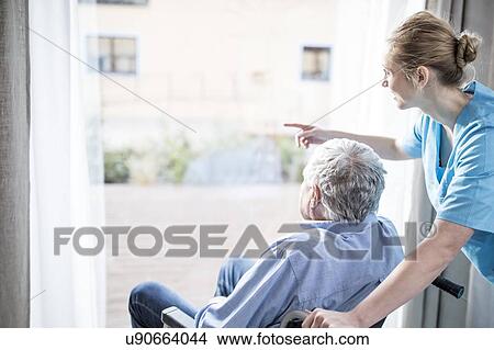 Picture - Senior man in wheelchair with care worker looking out of window.. Fotosearch