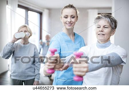 Stock Image - Senior woman holding hand weights, physiotherapist helping.. Fotosearch