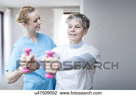 Stock Image - Senior woman holding hand weights, physiotherapist helping.. Fotosearch