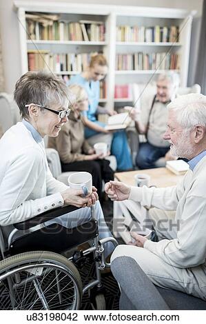 Stock Image - Senior woman in wheelchair talking to male fiend in care home.. Fotosearch