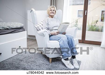 Stock Image - Senior woman sitting in armchair with magazine in care home.. Fotosearch