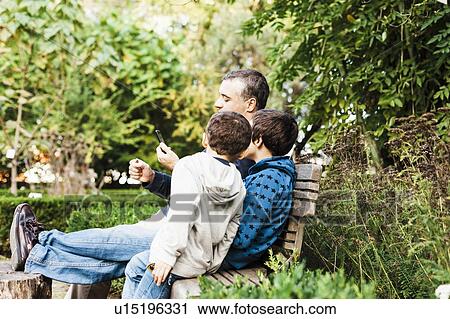 Stock Image - Father and two sons (10-11) sitting on bench in park and looking at smartphone. Fotosearch