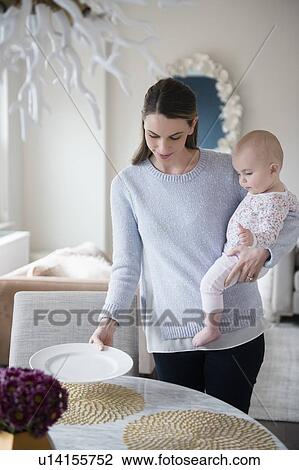 Stock Image - Mother with baby (18-23 months) in arms setting table. Fotosearch