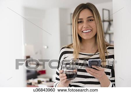 Stock Image - Portrait of young woman holding red wine and smart phone. Fotosearch