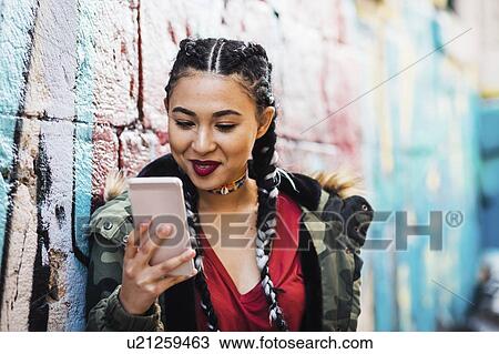 Stock Image - Portugal, Lisbon, Smiling young woman looking at smartphone, leaning against wall. Fotosearch