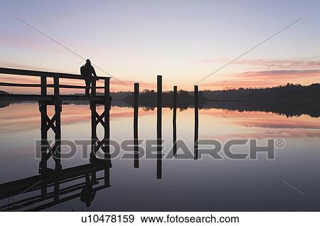 Stock Photo - Silhouette of man on pier at sunrise. Fotosearch