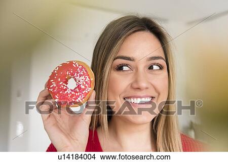 Picture - Smiling young woman holding donut with pink icing. Fotosearch