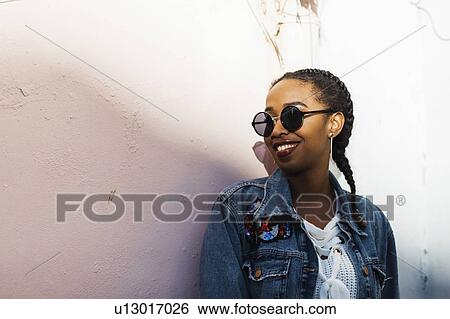 Stock Photograph - Smiling young woman wearing sunglasses and denim jacket standing by white wall. Fotosearch