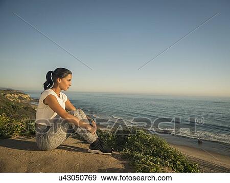 Stock Photo - USA, California, Newport Beach, Woman in sport suit sitting on cliff and looking at view. Fotosearch