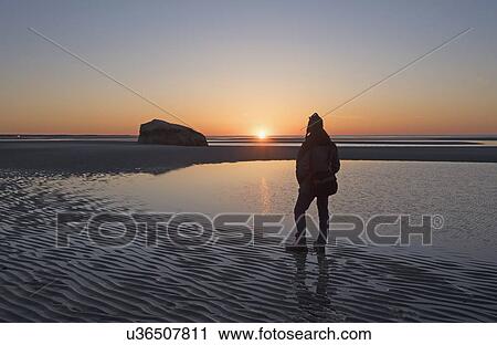 Stock Image - USA, Massachusetts, Cape Cod, Orleans, Man watching sunset at low tide. Fotosearch