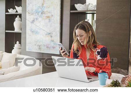 Stock Image - Woman using laptop and smart phone at home. Fotosearch