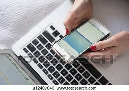 Stock Image - Woman with smartphone and laptop in bed. Fotosearch