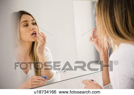 Stock Photograph - Young woman looking in mirror and applying lip balm. Fotosearch