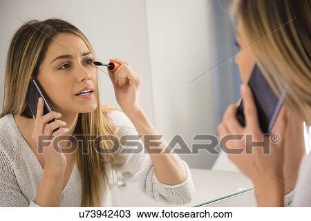 Stock Image - Young woman using smart phone and applying mascara. Fotosearch