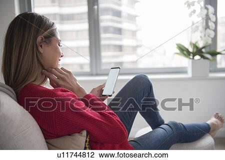 Stock Image - Young woman using smart phone in living room. Fotosearch
