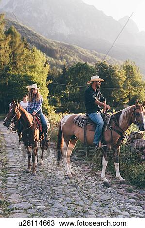 Stock Image - Adult friends horse riding on rural cobbled road, Primaluna, Trentino-Alto Adige, Italy. Fotosearch