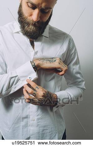 Stock Image - Bearded young man wearing white shirt, doing up cuff links, tattoos on hands. Fotosearch