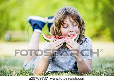 Stock Photo - Boy eating watermelon while lying in park, smiling, portrait.. Fotosearch