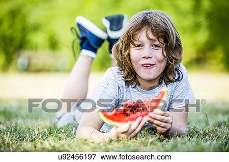 Stock Photo - Boy eating watermelon while lying in park, smiling, portrait.. Fotosearch