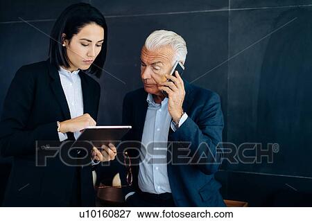 Stock Photo - Businessman and woman looking at digital tablet and making smartphone call in boardroom. Fotosearch