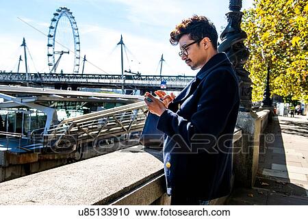 Stock Image - Businessman using smartphone on Embankment, London Eye and Hungerford Bridge in background, London, UK. Fotosearch