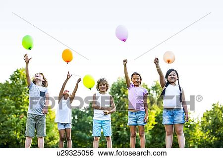 Stock Photograph - Children standing in a row with balloons in park, smiling.. Fotosearch