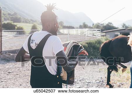 Stock Photo - Cool young man carrying saddle in rural equestrian arena. Fotosearch