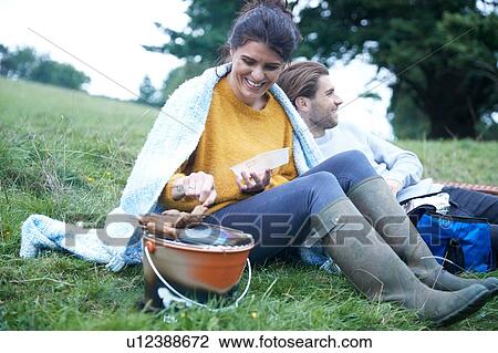 Stock Image - Couple sitting in rural field cooking on barbecue grill. Fotosearch