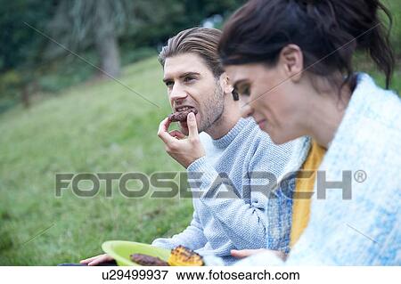 Stock Photo - Couple sitting in rural field eating barbecue food. Fotosearch