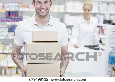 Stock Image - Courier carrying boxes in pharmacy with pharmacist in background.. Fotosearch