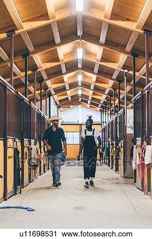 Stock Image - Cowboy and cool young man walking in stable block. Fotosearch