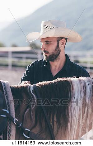 Stock Photo - Cowboy checking horse bridle in equestrian arena, cropped. Fotosearch