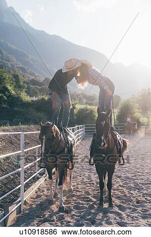 Stock Photograph - Cowgirl and boyfriend standing on horseback kissing in equestrian arena, Primaluna, Trentino-Alto Adige, Italy. Fotosearch