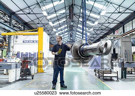 Stock Image - Engineer moving gear and spindle on crane in gearbox factory. Fotosearch