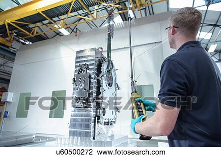 Stock Image - Engineer using crane to lift parts onto CNC machine. Fotosearch