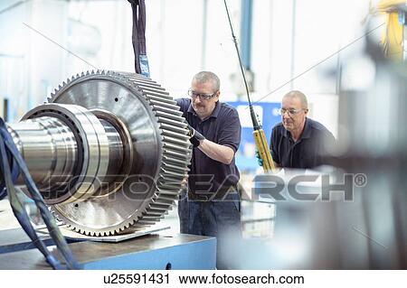 Stock Image - Engineers using crane to move large gear in gearbox factory. Fotosearch