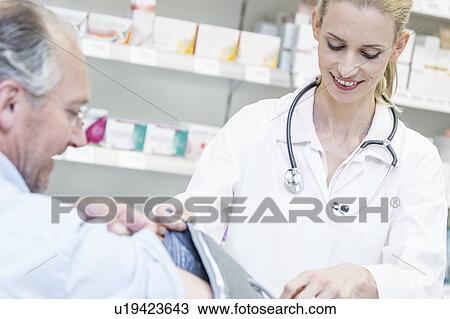 Stock Image - Female pharmacist checking mature male patient's blood pressure in pharmacy.. Fotosearch