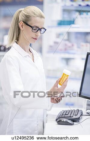 Stock Photograph - Female pharmacist reading label on pill bottle in pharmacy.. Fotosearch