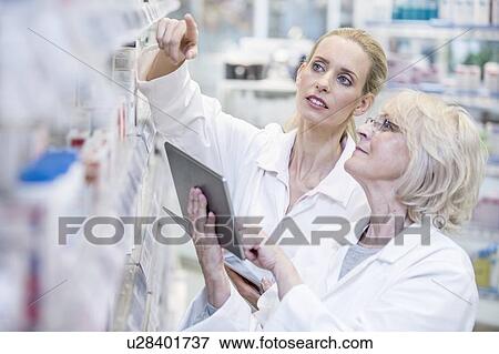 Stock Photo - Female pharmacists checking medicines on shelf using digital tablet.. Fotosearch