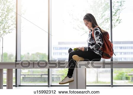Stock Image - Female university student sitting on table reading a book in university lobby. Fotosearch