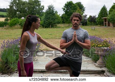 Stock Image - Female yoga teacher teaching young man yoga in garden, tree pose. Fotosearch